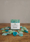 A rectangular bar of TARTME & COMPANY Sauvage soap with a white label and green swirls, displayed on a wooden surface with scattered blue and green petals against a neutral wall.